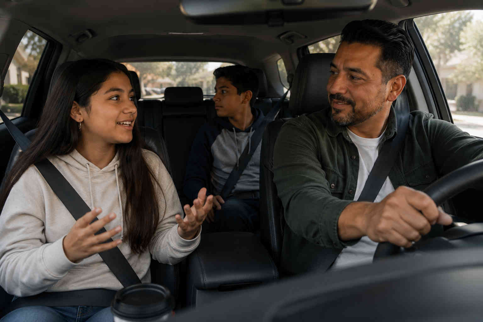 Father driving while his teenage daughter talks comfortably and his son sits in the back seat during a ride between homes