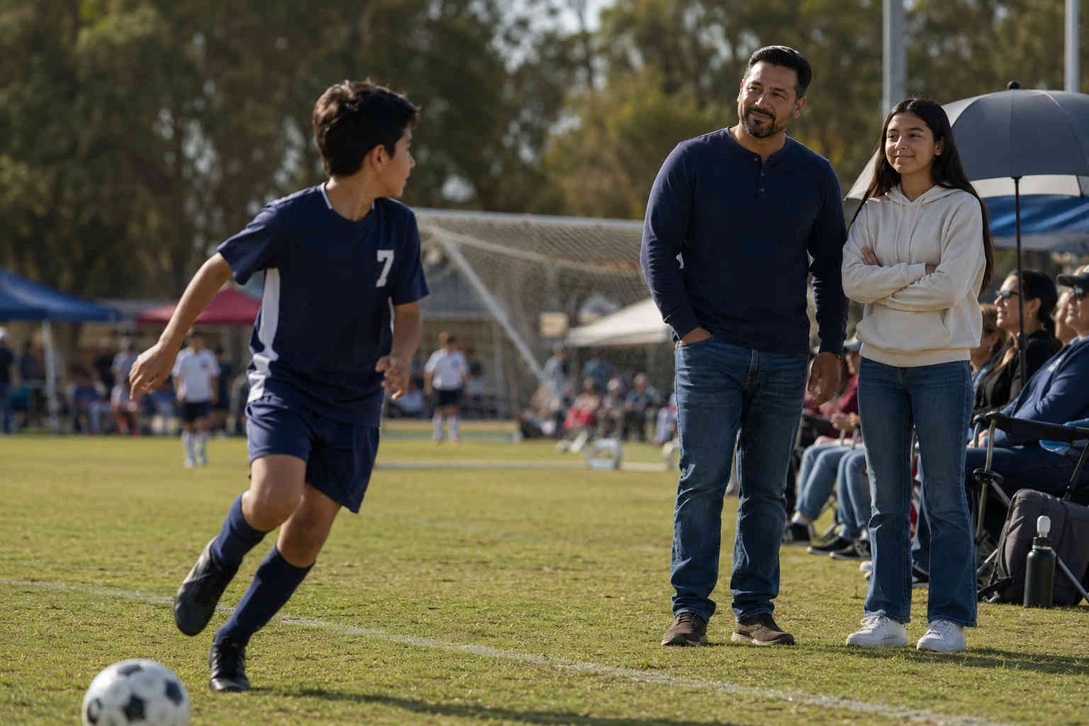 Father and teenage daughter standing on the sideline as his son looks toward them during a youth soccer game